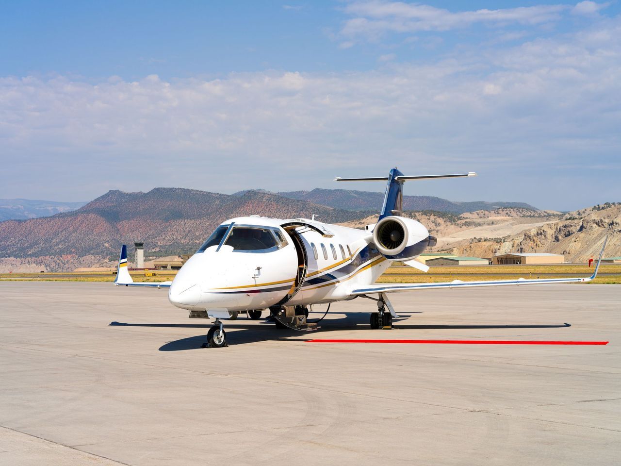 Jet Charter A small white private jet with an open door is parked on a sunny airport runway, ready for exclusive flights from New York to Los Angeles, with mountains and a partly cloudy sky in the background. Jetvia