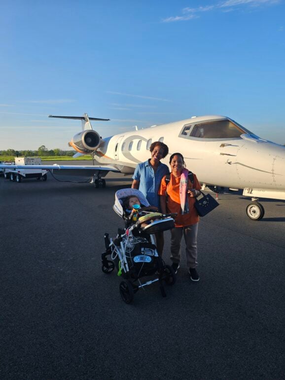 Jet Charter Two adults and a baby in a stroller stand on an airport tarmac in front of a small private jet under a clear blue sky. The adults smile at the camera. Jetvia