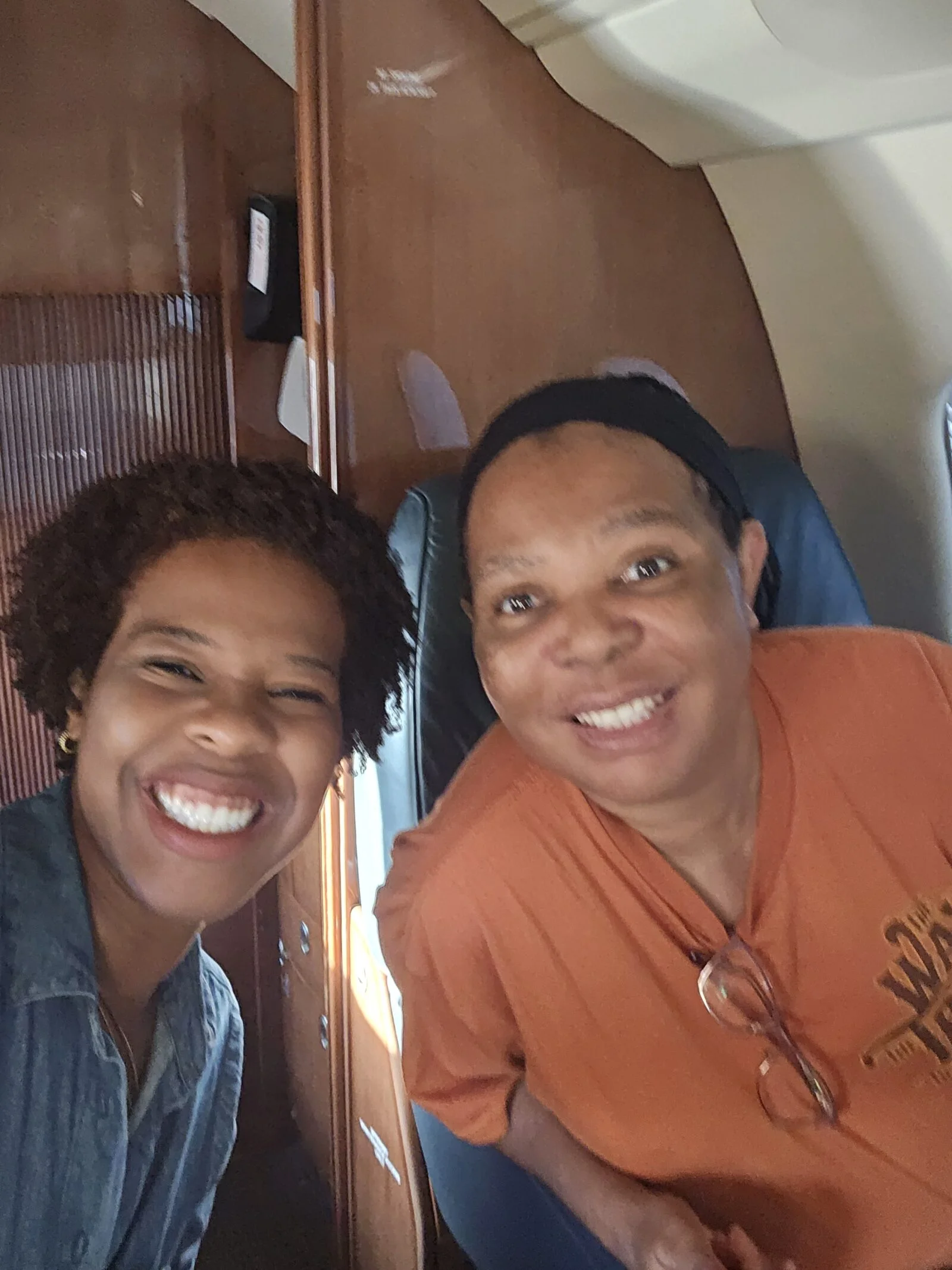 Jet Charter Two people smiling and posing for a selfie inside an airplane, with wood paneling and blue seats visible in the background. Jetvia