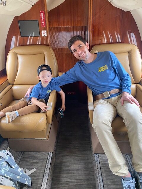 Jet Charter A man and a young boy, both in blue shirts and khaki pants, smile while seated in tan leather seats on a private jet. The boy wears a backwards cap and holds a toy car. Jetvia