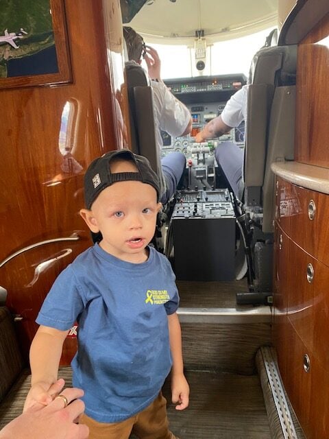 Jet Charter A young boy in a blue shirt and backwards cap stands inside an airplane cabin, near the cockpit, holding an adult’s hand. Two pilots are visible seated at the controls in the background. Jetvia