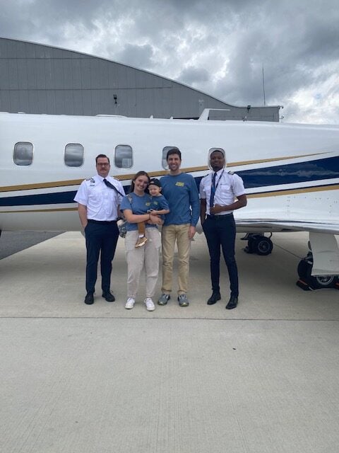 Jet Charter Four adults and one child stand smiling in front of a small white jet on an airport tarmac. Two adults wear pilot uniforms, while the others are in casual clothing. A hangar and cloudy sky are visible in the background. Jetvia