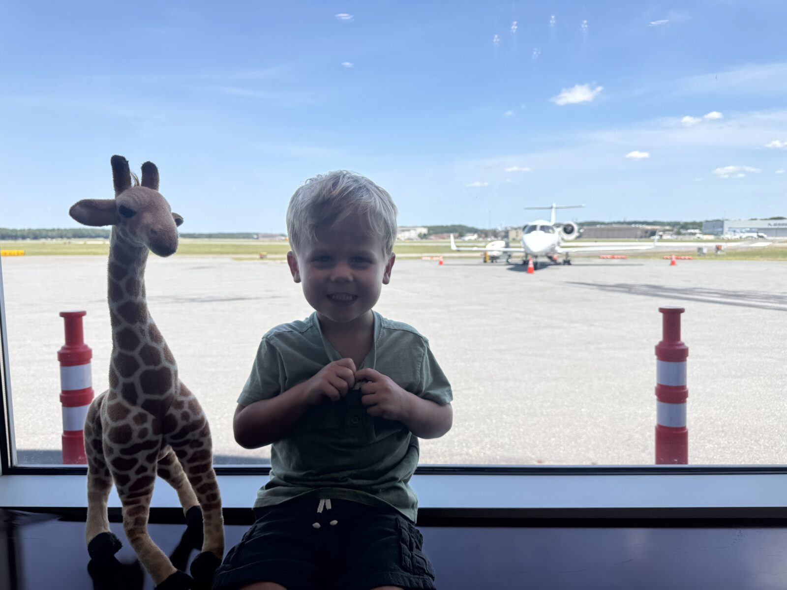 Jet Charter A young boy sits next to a stuffed giraffe by an airport window, smiling, with a small jet and blue sky visible outside. Jetvia