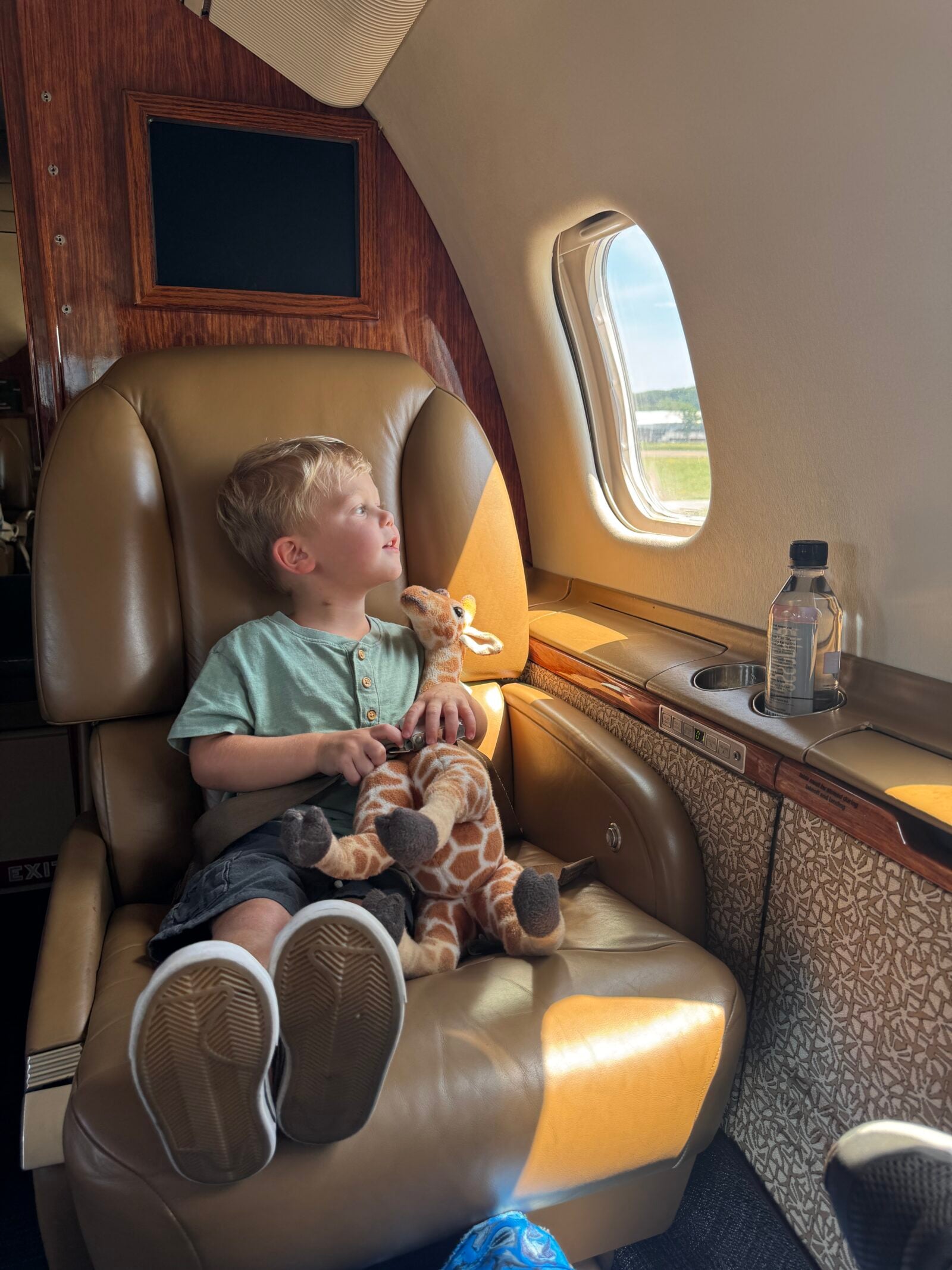 Jet Charter A young boy sits in a leather airplane seat, holding a stuffed giraffe and looking out the window. A bottle of water is on the ledge beside him. Sunlight shines on his face. Jetvia