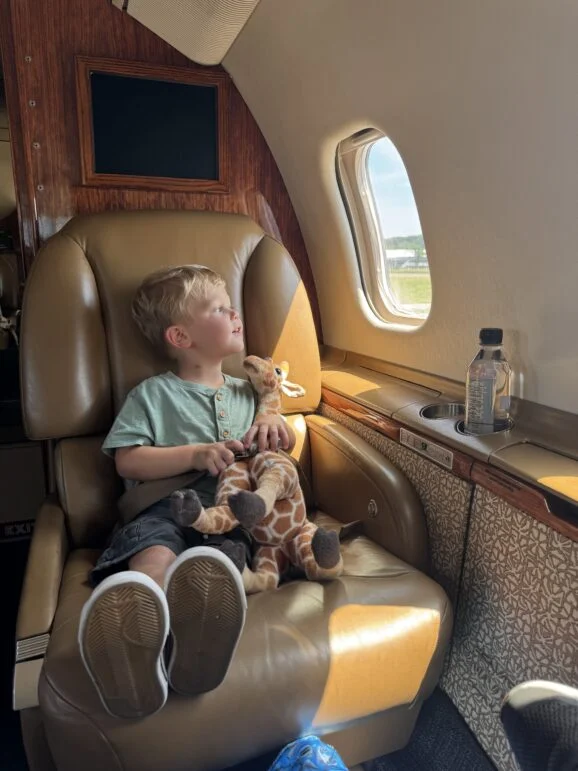 Jet Charter A young boy sits in a leather airplane seat, holding a stuffed giraffe and looking out the window. A bottle of water is on the ledge beside him. Sunlight shines on his face. Jetvia