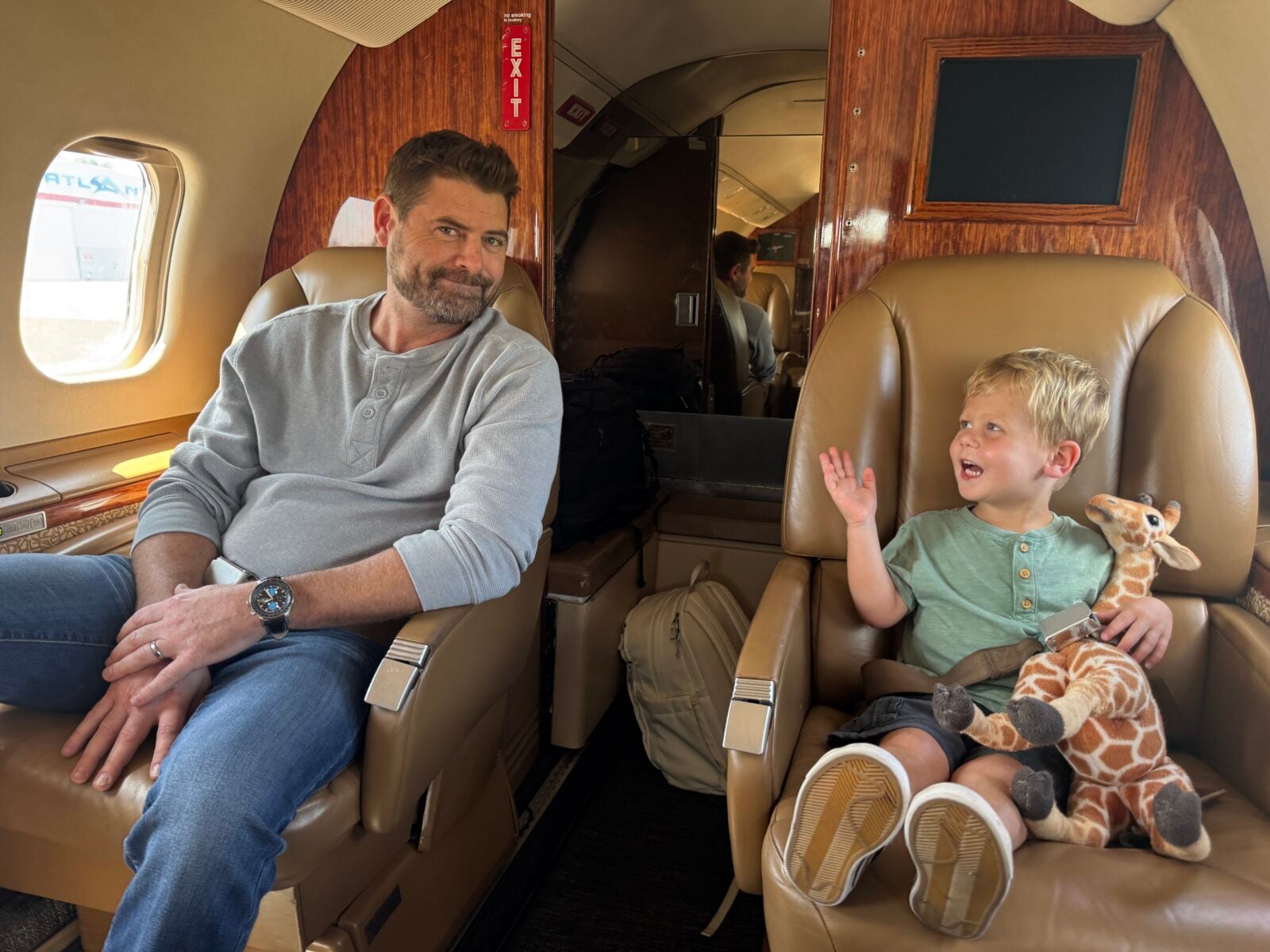 Jet Charter A man and a young boy sit in tan leather seats on a private jet. The boy, holding a stuffed giraffe, gestures while smiling at the man, who looks at him with a slight grin. Jetvia