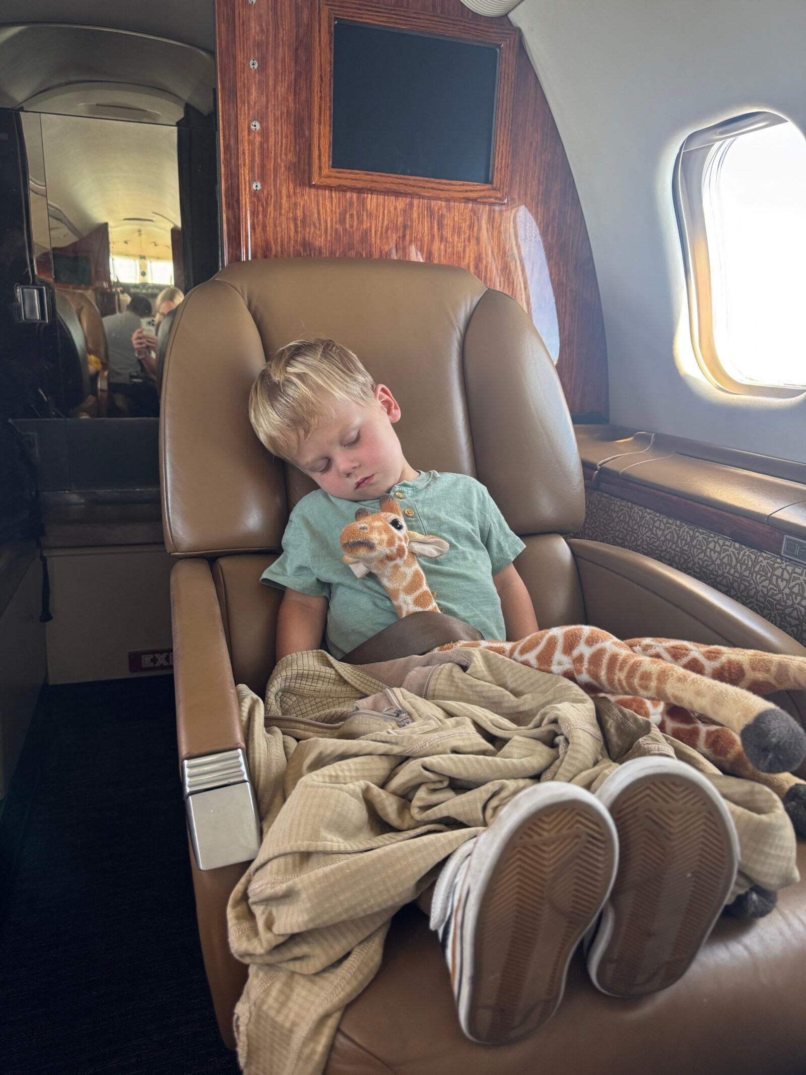 Jet Charter A young boy sleeps in a leather airplane seat, cuddling a small giraffe plush toy. A beige blanket covers him and the seat. Sunlight streams through the window beside him. Jetvia