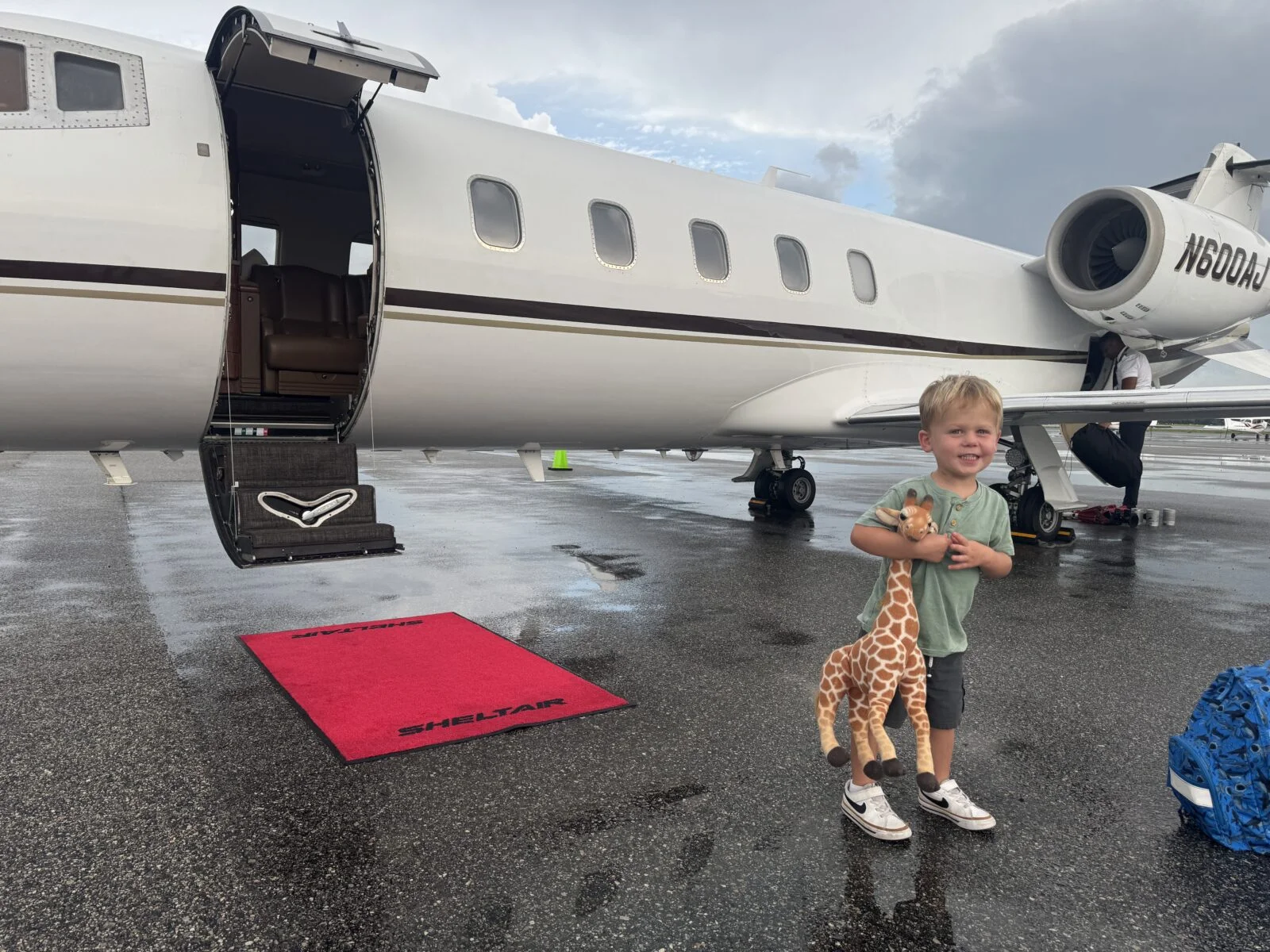 Jet Charter A young boy holding a toy giraffe stands near a private jet on a wet tarmac, with the jet’s door open and a red carpet rolled out. Jetvia