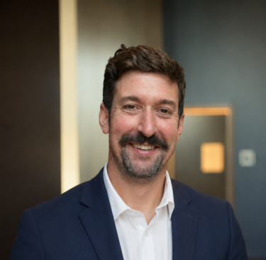 Jet Charter A man with dark hair and a mustache, wearing a navy suit and white shirt, smiles while standing indoors with a blurred background. Jetvia