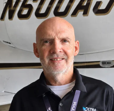 Jet Charter A man with a shaved head wearing a black JetWAV polo stands in front of a white jet with the tail number N600AJ inside an aircraft hangar. This is Scotty Woodall. Jetvia