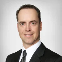 Jet Charter A man in a black suit, white shirt, and patterned tie smiles at the camera against a plain, light gray background. Jetvia