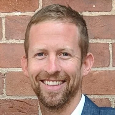 Jet Charter A smiling man with short light brown hair and a beard stands in front of a red brick wall, wearing a blue jacket and white shirt. Jetvia
