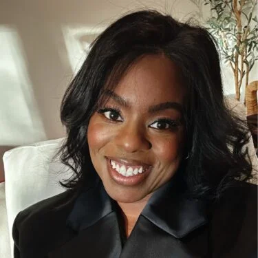 Jet Charter Stevie Coleman, a woman with shoulder-length black hair and a black top, smiles at the camera while sitting indoors, with sunlight and a plant in the background. Jetvia