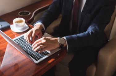 Jet Charter A person in a suit types on a laptop on a wooden tray table next to a cup of coffee. Jetvia